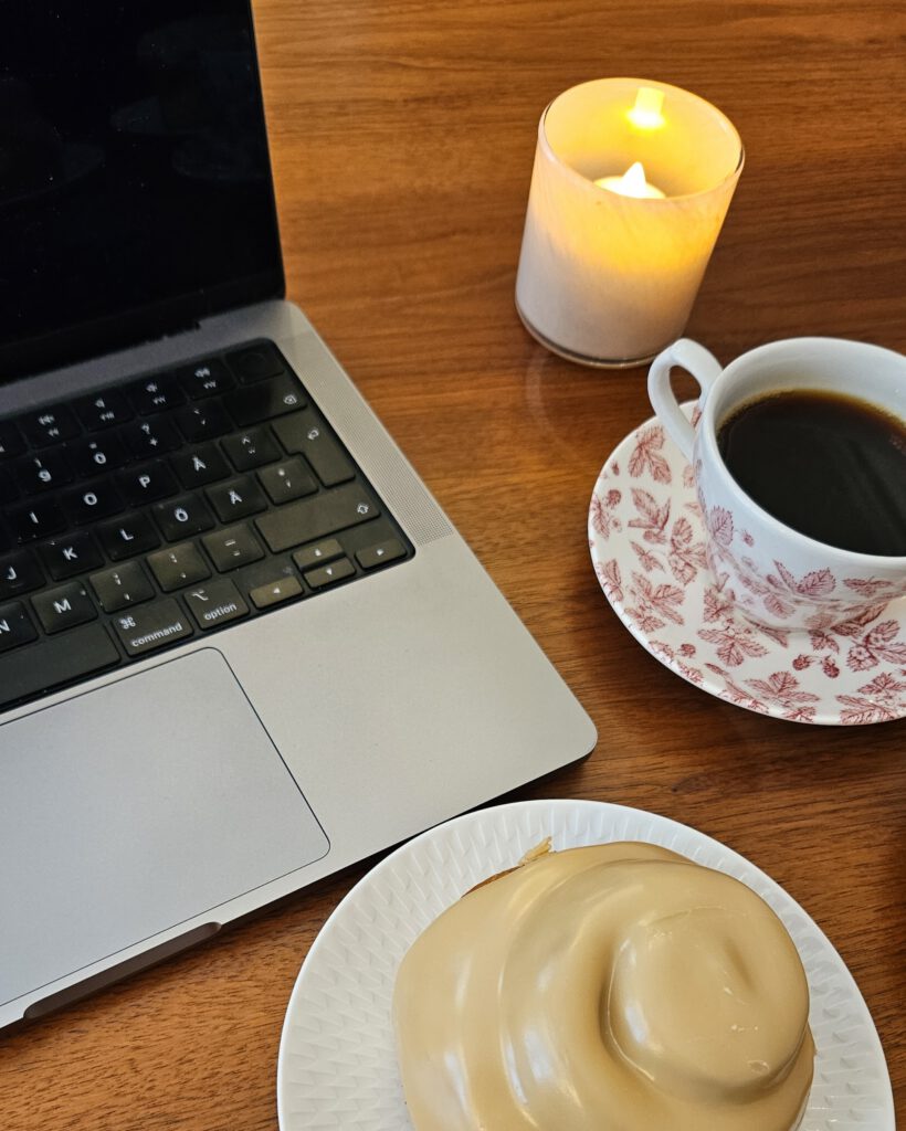 Pöydällä Macbook, pulla, kuppi kahvia ja kynttilä.