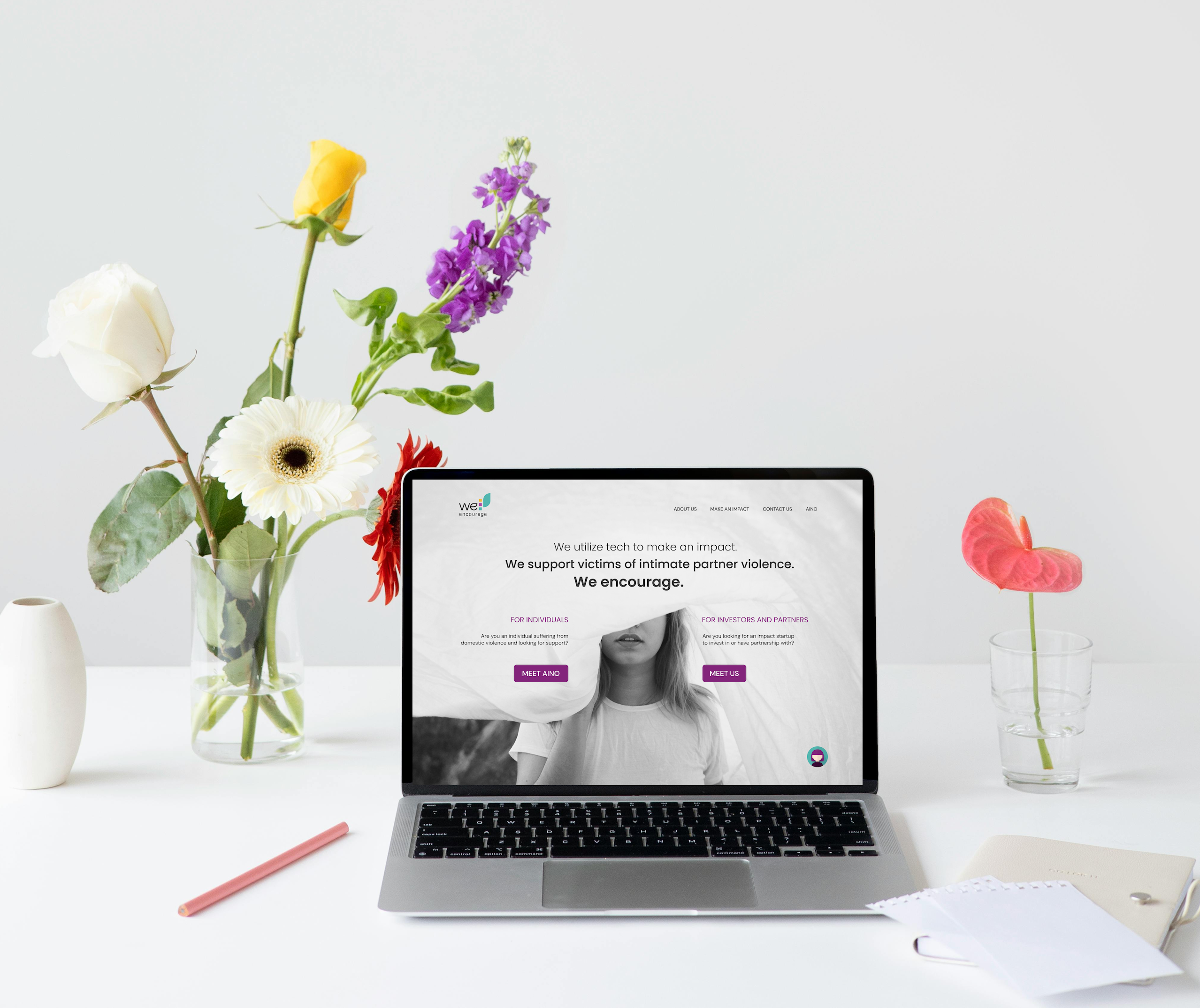 We Encourage -sivuston etusivu Macbookin näytöllä. Macbookin lisäksi valkoisella pöydällä on kukkia maljakossa ja vesilasissa, muistikirja sekä kyniä.